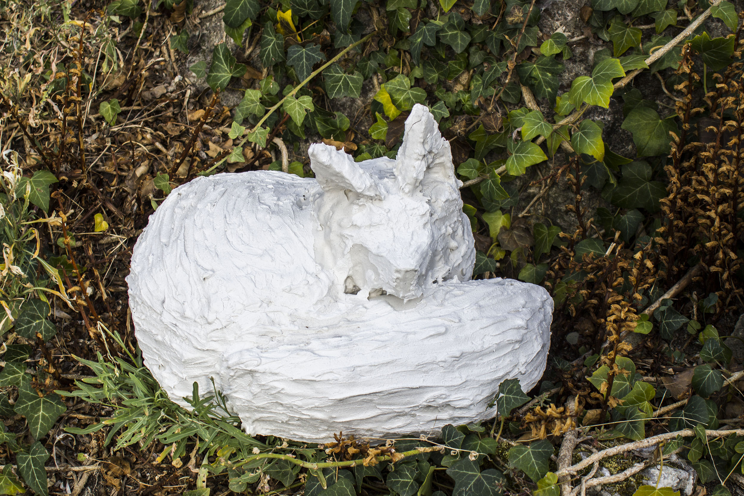 Une sculpture de renardeau lové, en plâtre blanc effet crépi est posé au milieu de lierre.